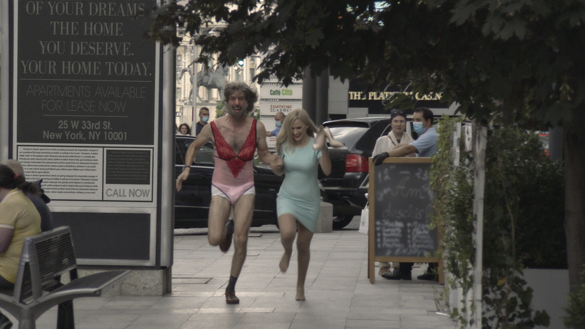 Borat and Tutar running down a sidewalk in a big city (Borat is wearing women's lingerie, Tutar is wearing a dress)