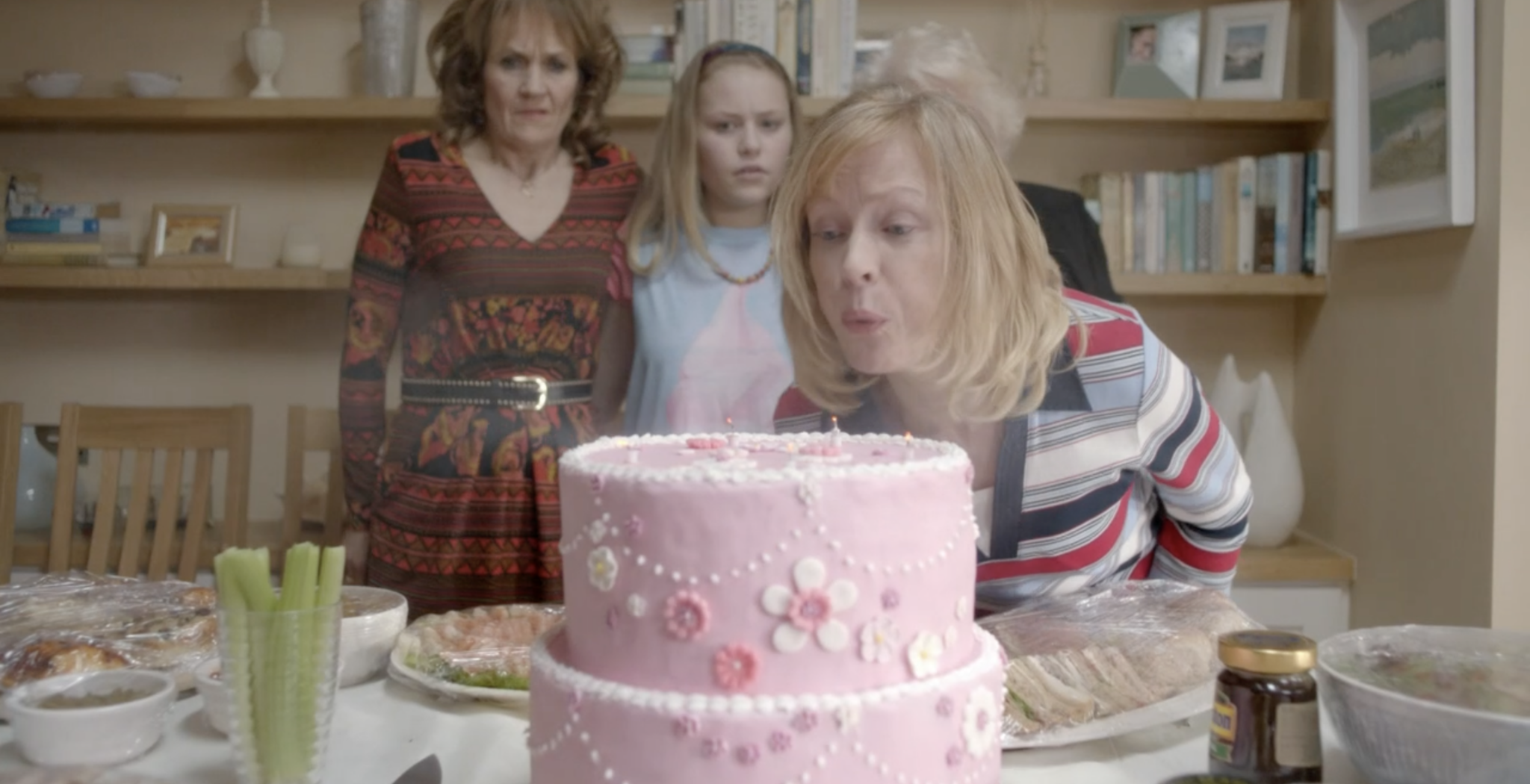 Angela blows out the candles on a pink birthday cake while everyone looks on, alarmed.